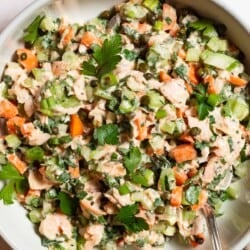 A overhead close up photo of salmon salad in a serving bowl with a spoon next to bowls of capers and parsley.