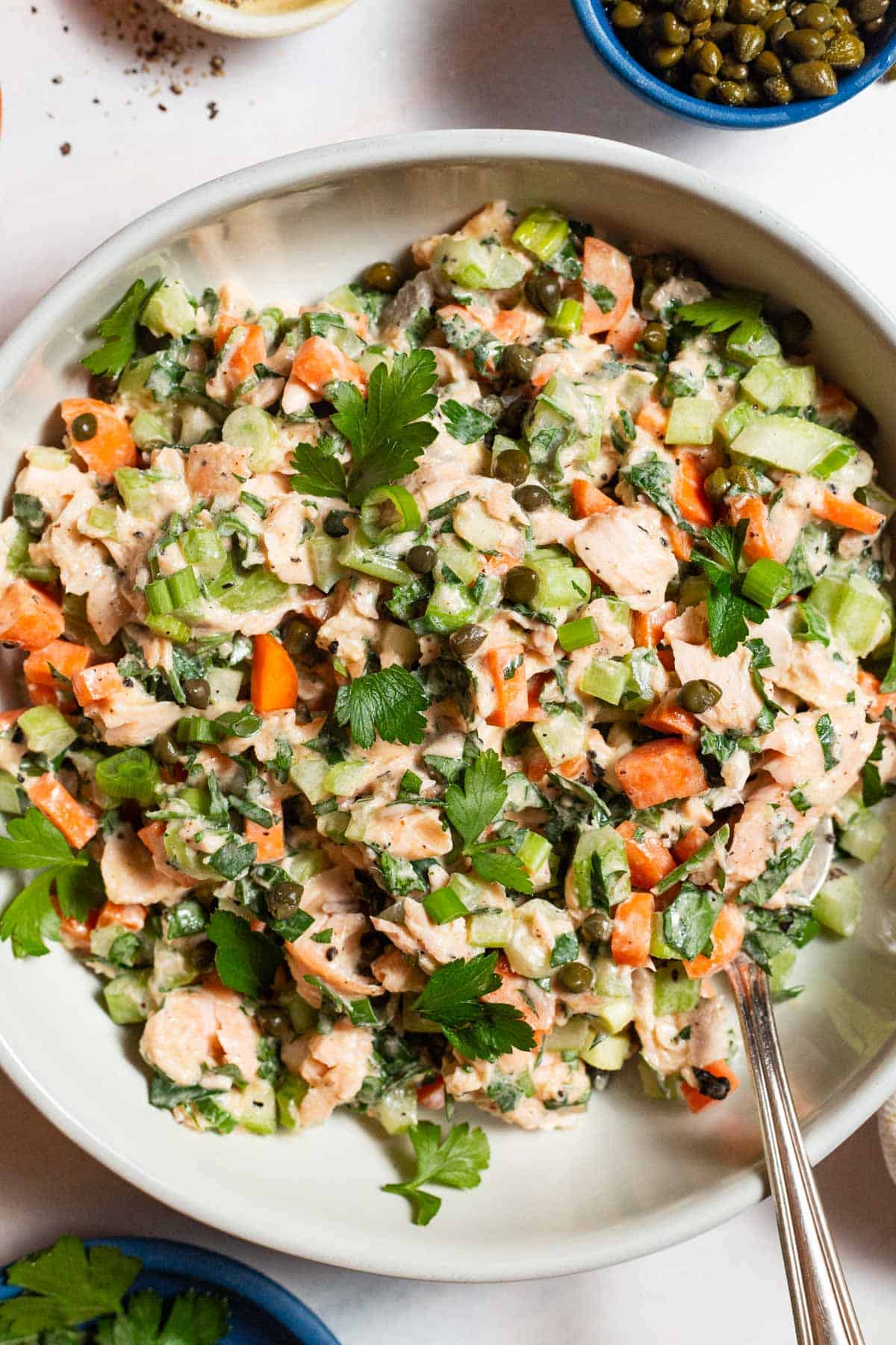 A overhead close up photo of salmon salad in a serving bowl with a spoon next to bowls of capers and parsley.