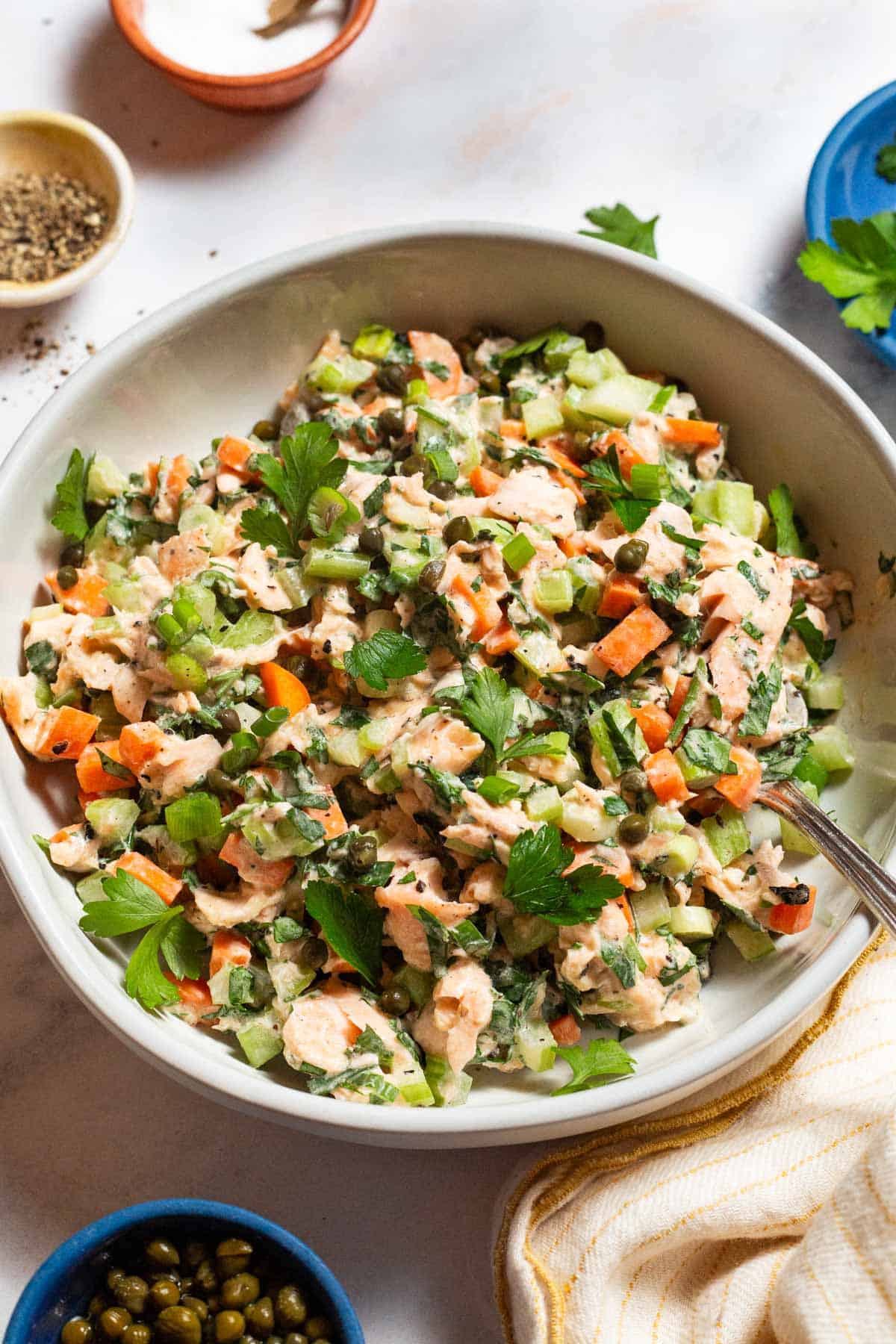 Salmon salad in a serving bowl with a spoon. Next to this are bowls of salt, pepper and capers, and a kitchen towel.