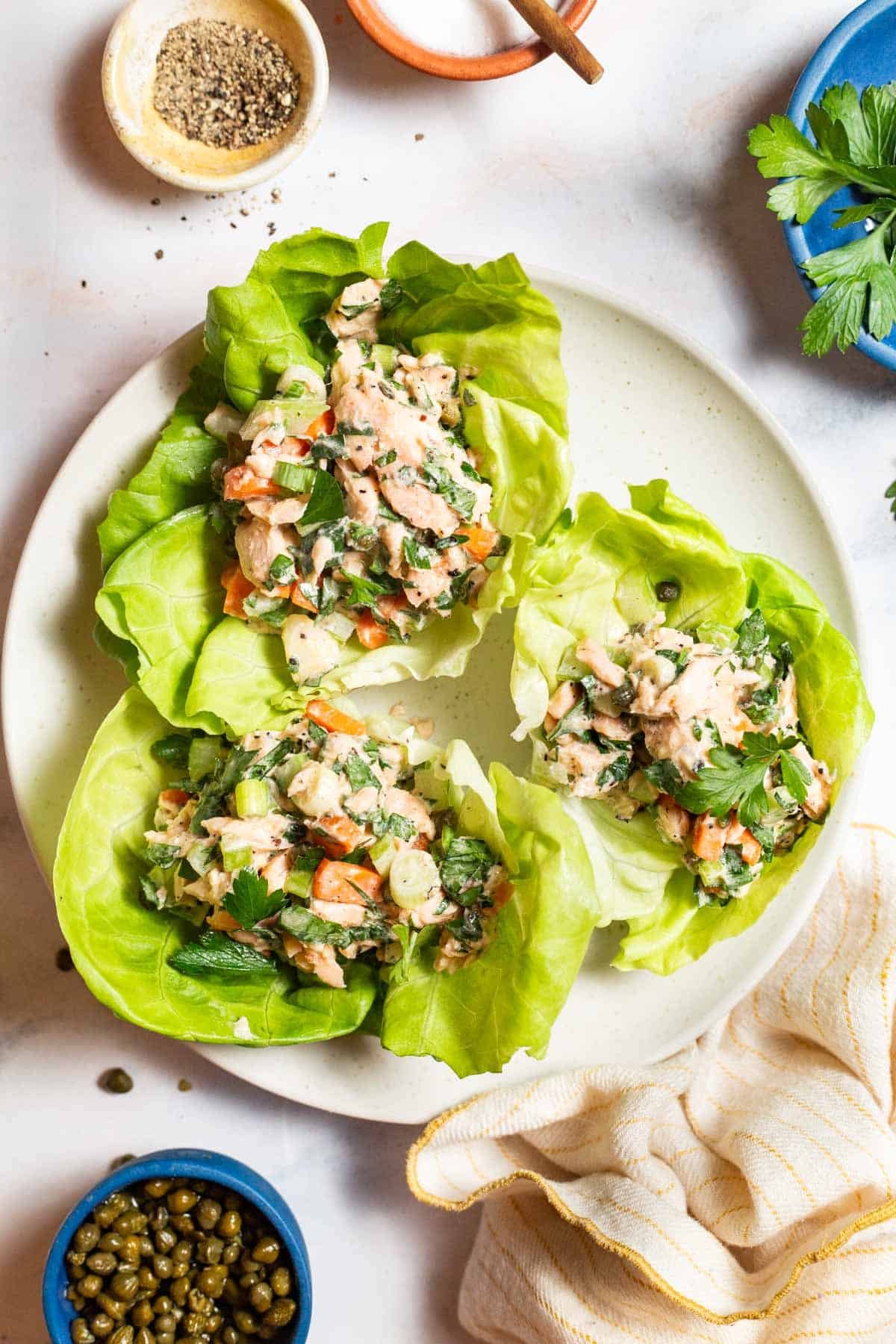 An overhead photo of 3 servings of the salmon salad in lettuce wraps on a plate surrounded by bowls of salt, pepper, capers and parsley and a kitchen towel.