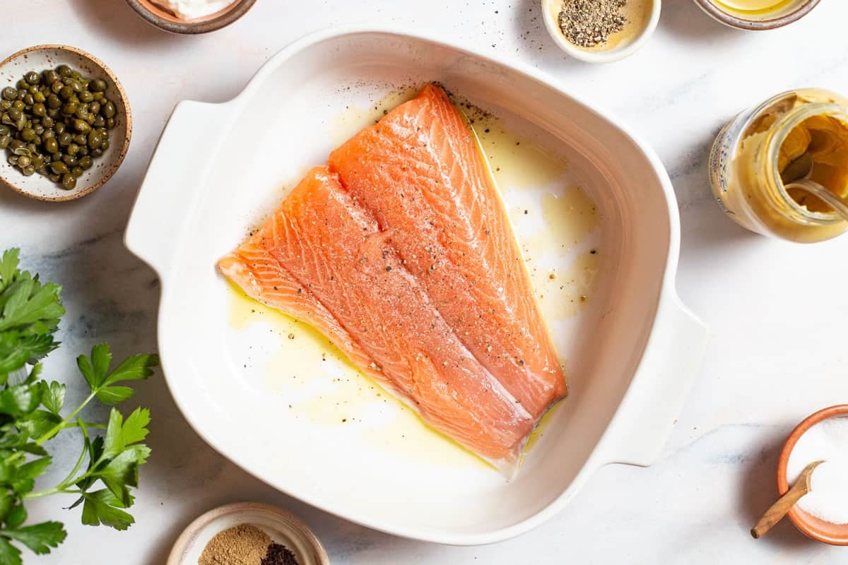 An uncooked salmon fillet in a baking dish. Next to this is parsley, a jar of dijon mustard and bowls of spices, salt, pepper and capers.