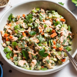 A close up of salmon salad in a serving bowl with a spoon next to bowls of pepper, parsley and capers, and a kitchen towel.