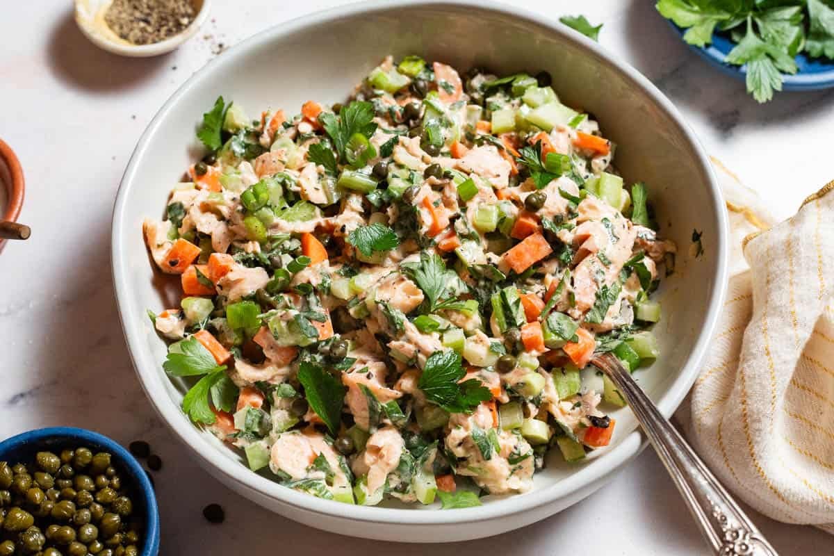 A close up of salmon salad in a serving bowl with a spoon next to bowls of pepper, parsley and capers, and a kitchen towel.