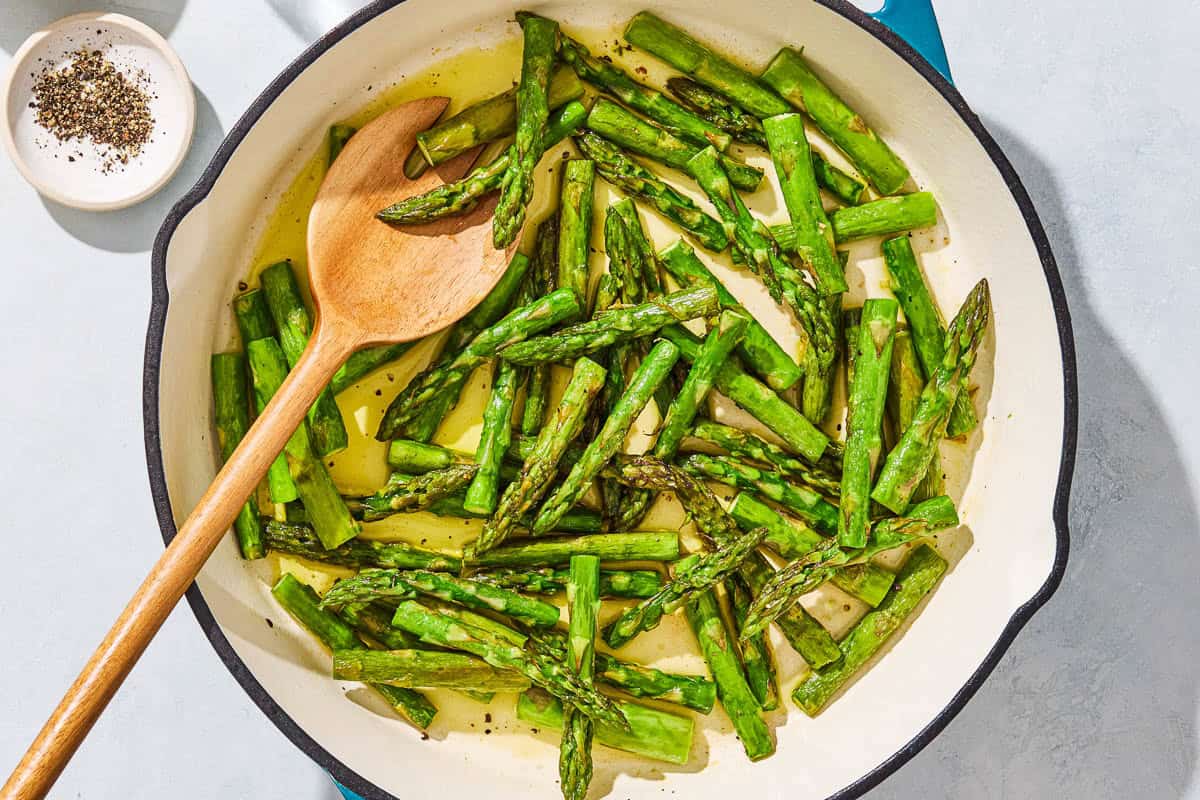 Sauteed asparagus in a skillet with a wooden fork next to a small bowl of pepper.