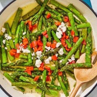 Sauteed asparagus topped with chopped red peppers and crumbled feta in a skillet with a wooden fork. Next to this is a bowl of crumbled feta.