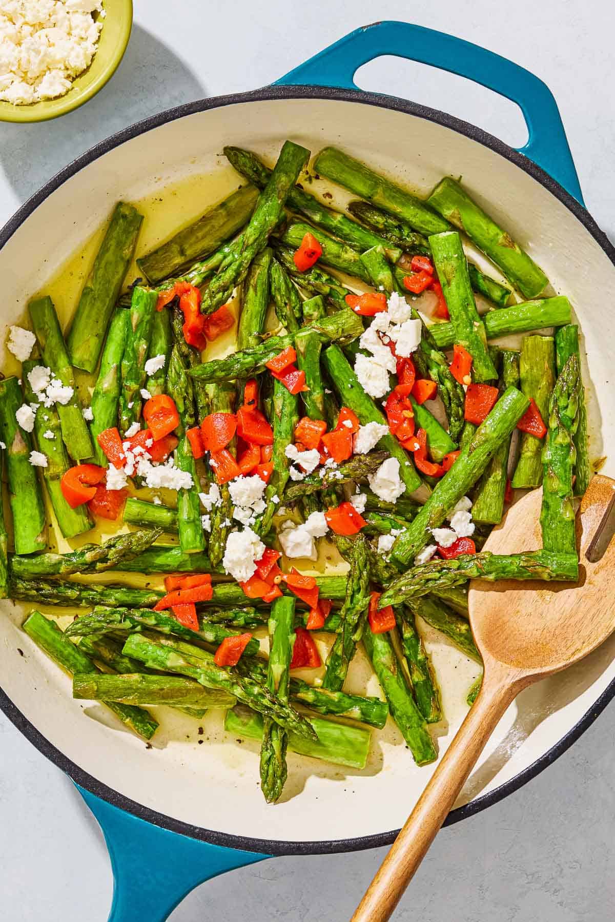 Sauteed asparagus topped with chopped red peppers and crumbled feta in a skillet with a wooden fork. Next to this is a bowl of crumbled feta.