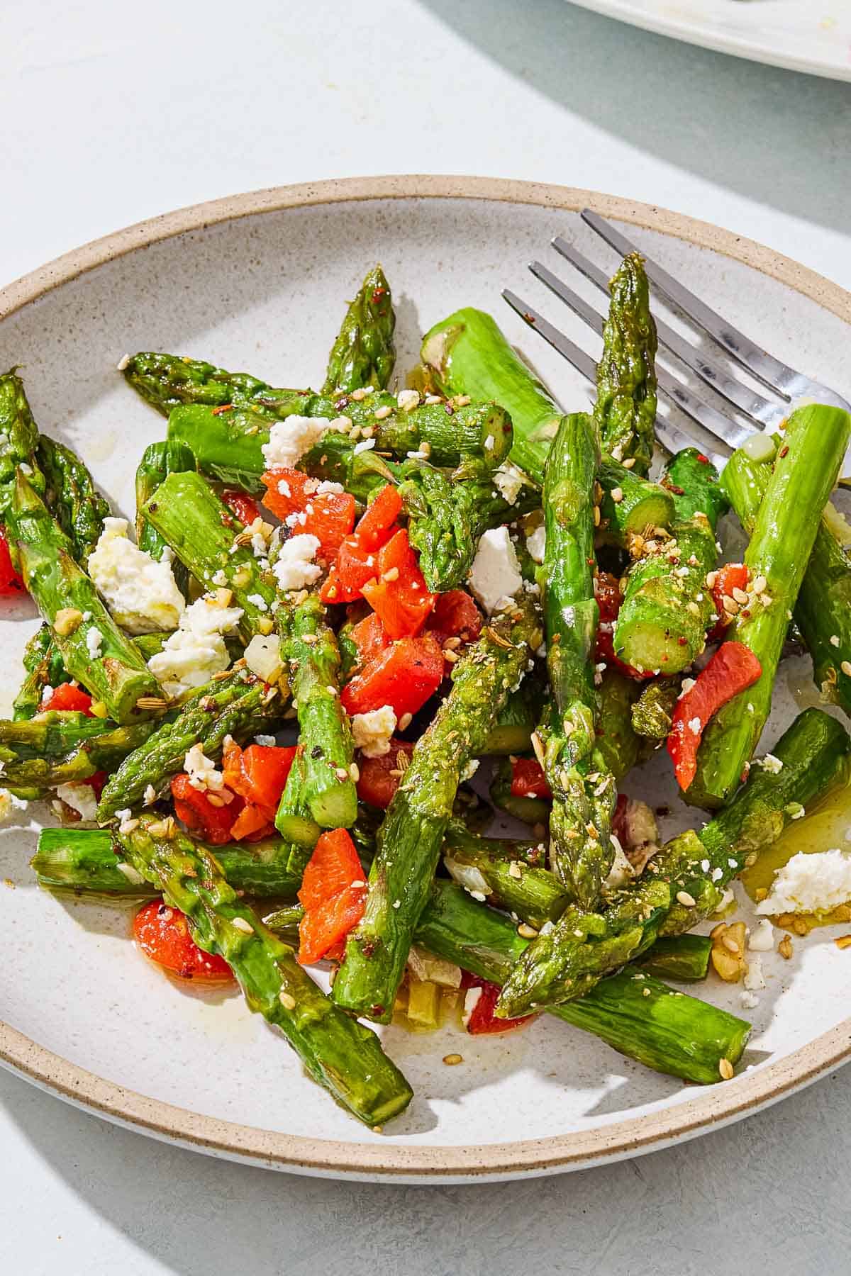 A close up of a serving of sauteed asparagus topped with chopped red peppers and crumbled feta on a plate with a fork.