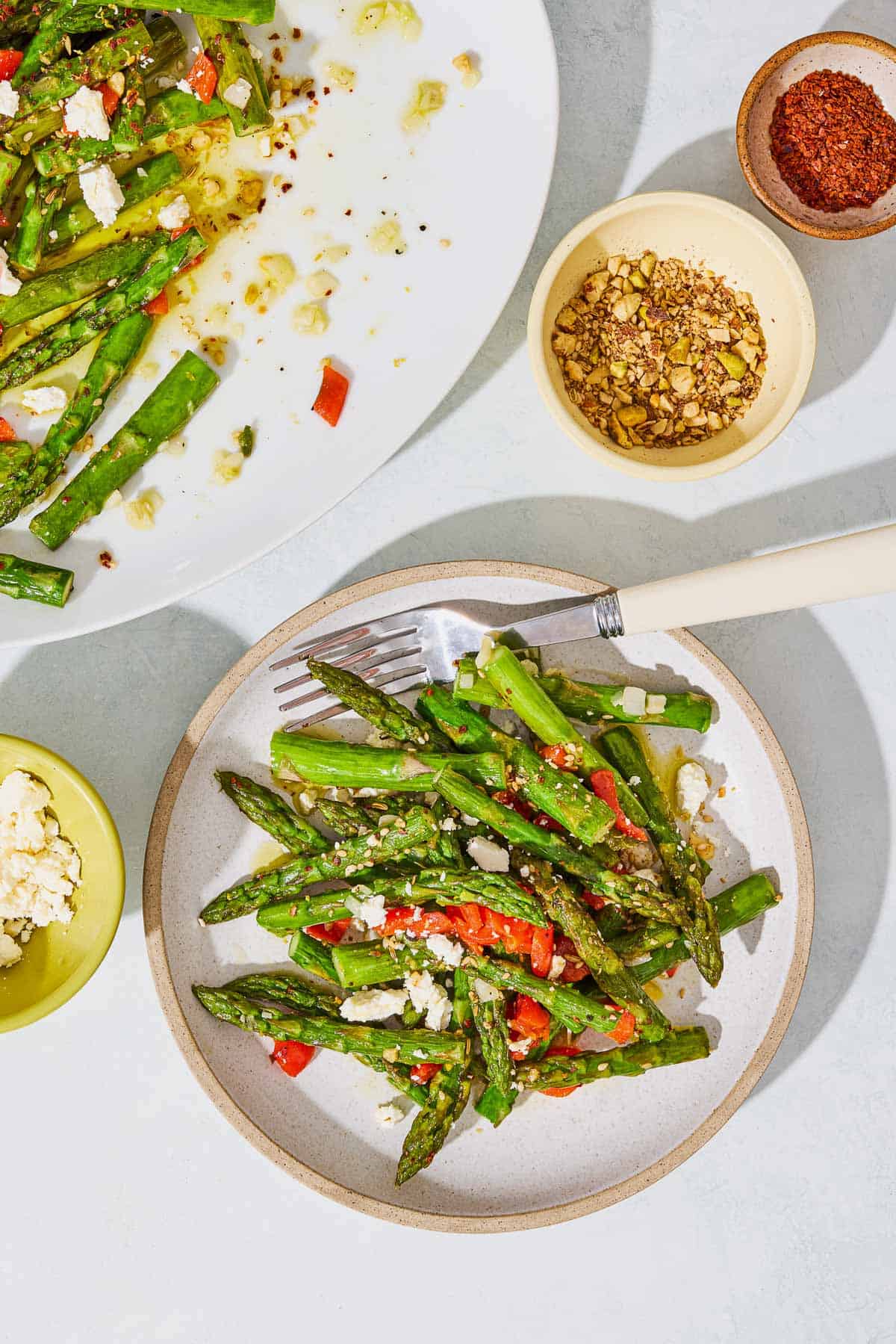 An overhead photo of a serving of sauteed asparagus topped with chopped red peppers and crumbled feta on a plate with a fork. Next to this is a platter with the rest of the asparagus and small bowls of crumbled feta, za'atar and aleppo pepper.