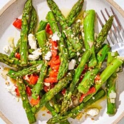 A serving of sauteed asparagus topped with chopped red peppers and crumbled feta on a plate with a fork.