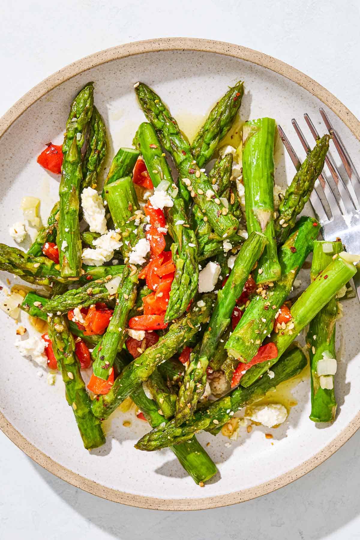A serving of sauteed asparagus topped with chopped red peppers and crumbled feta on a plate with a fork.
