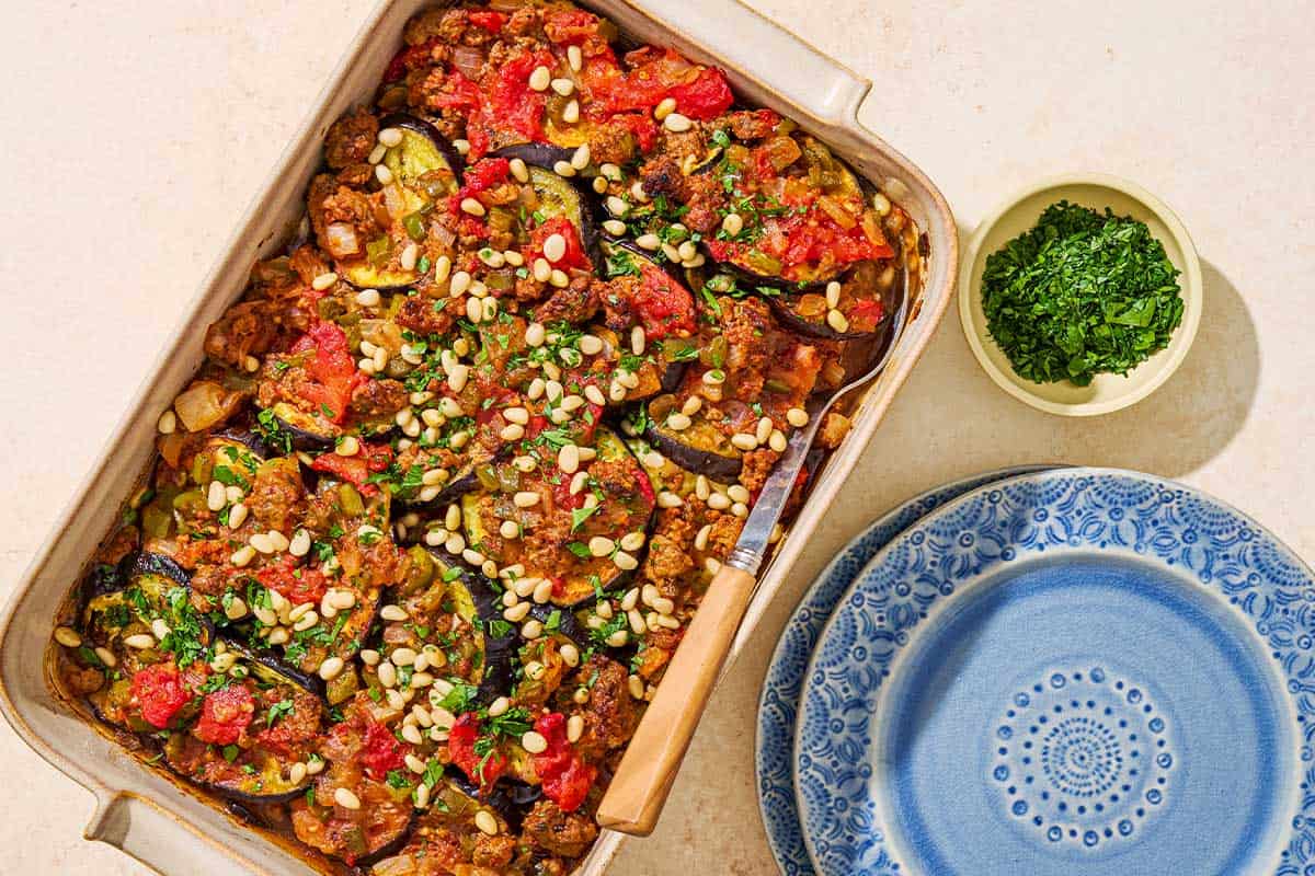 An overhead photo of egyptian moussaka in a baking dish with a serving spoon next to a stack of 2 plates and a bowl of chopped parsley.