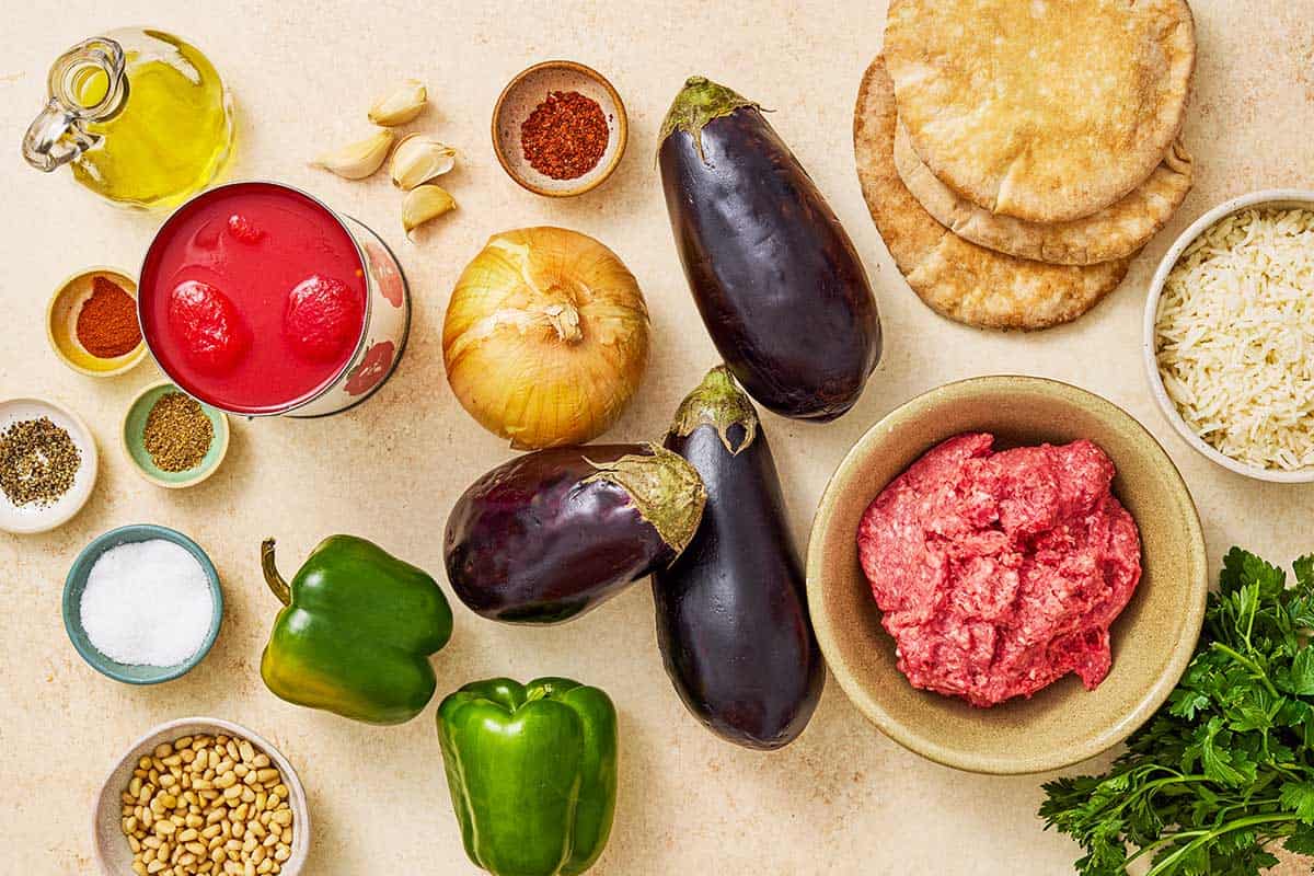 ingredients for Egyptian moussaka including eggplant, salt, onion, green bell peppers, garlic, olive oil, ground beef, coriander, sweet paprika, aleppo pepper, black pepper, san marzano tomatoes, parsley, pine nuts, and pita bread.