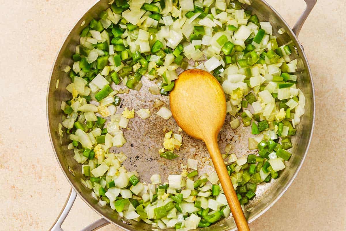Chopped onion and green peppers being sauteed in a skillet with a wooden spoon.
