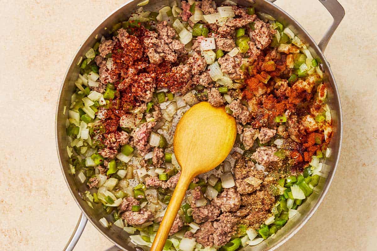 Ground beef, chopped onion, chopped green peppers and spices being sauteed in a skillet with a wooden spoon.