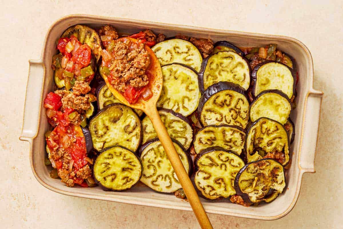 Meat sauced being added to a layer of sliced, roasted eggplant in a baking dish with a wooden spoon.