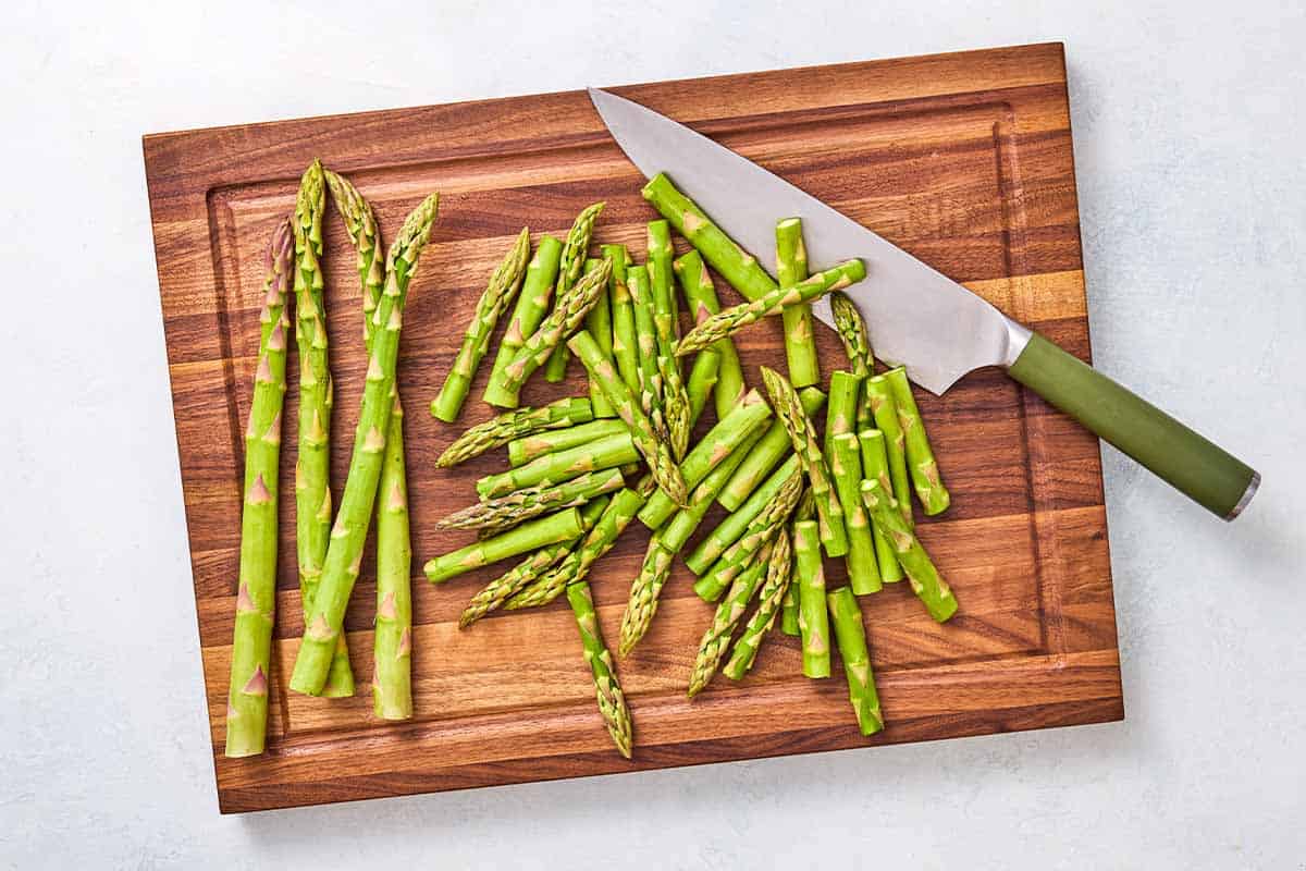 Cut up pieces of asparagus on a cutting board with pieces of whole asparagus and a knife.