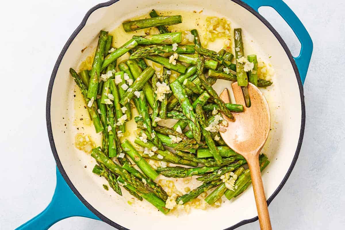 Asparagus being sauteed with garlic in a skillet with a wooden fork.