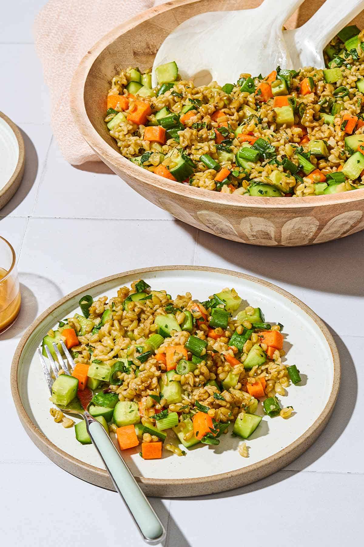 The herby freekeh salad in a serving bowl with serving utensils next to a serving of the salad on a plate with a fork.