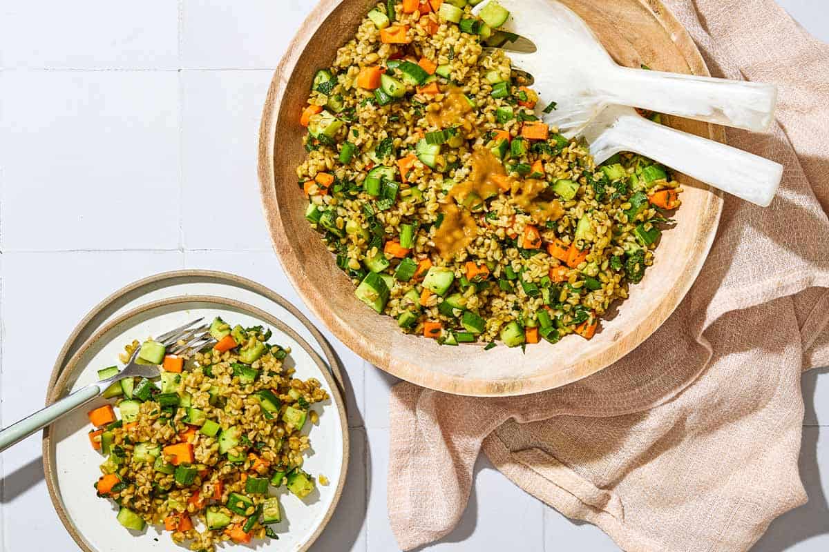An overhead photo of herby freekeh salad in a serving bowl with serving utensils next to a kitchen towel and a serving of the salad on a plate with a fork.