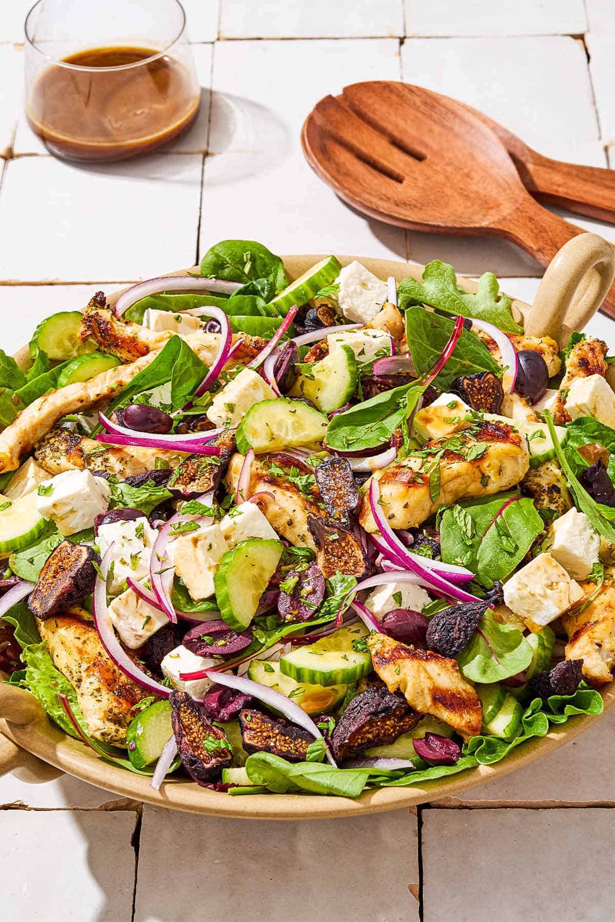 A Greek-stye chicken spinach salad in a serving bowl next to wooden serving utensils and a glass of the dressing.