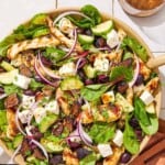 An overhead photo of Greek-stye chicken spinach salad in a serving bowl next to wooden serving utensils and a glass of the dressing.