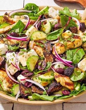 An overhead photo of Greek-stye chicken spinach salad in a serving bowl next to wooden serving utensils and a glass of the dressing.