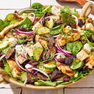 An overhead photo of Greek-stye chicken spinach salad in a serving bowl next to wooden serving utensils and a glass of the dressing.