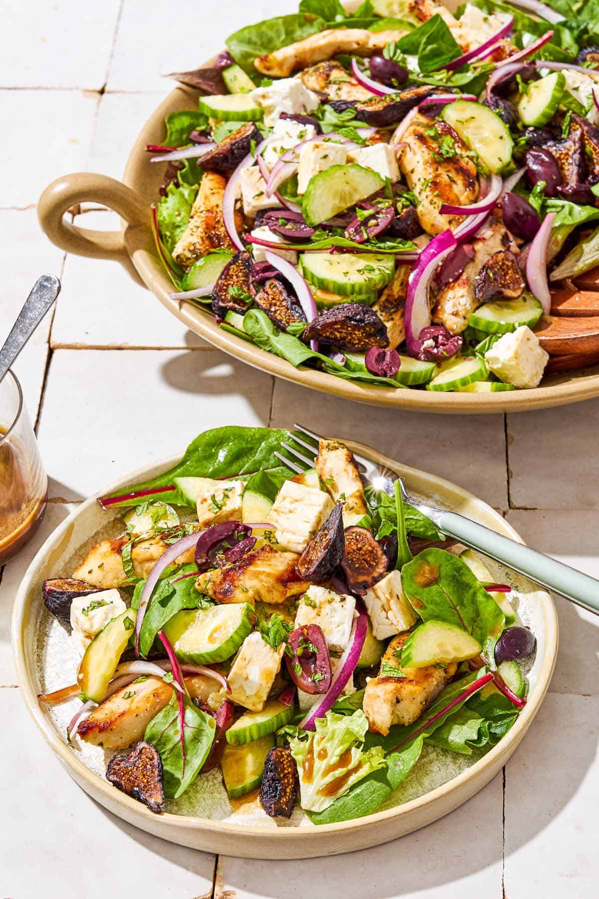 A Greek-stye chicken spinach salad in a serving bowl next to a plate of the salad with a fork.