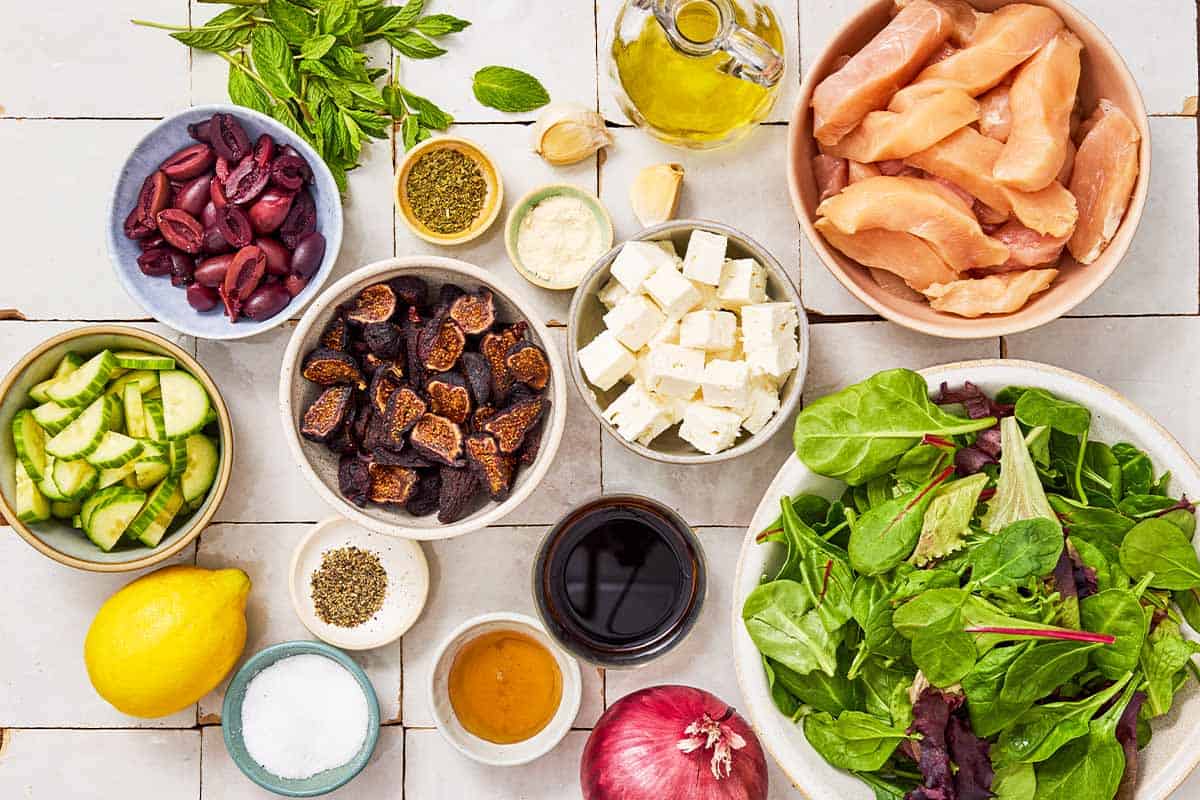 Ingredients for Greek-stye chicken spinach salad including chicken breasts, olive oil, oregano, garlic, onion powder, lemon, salt black pepper, balsamic vinegar, honey, baby spinach, red onion, cucumber, kalamata olive, dried figs, feta, and mint.