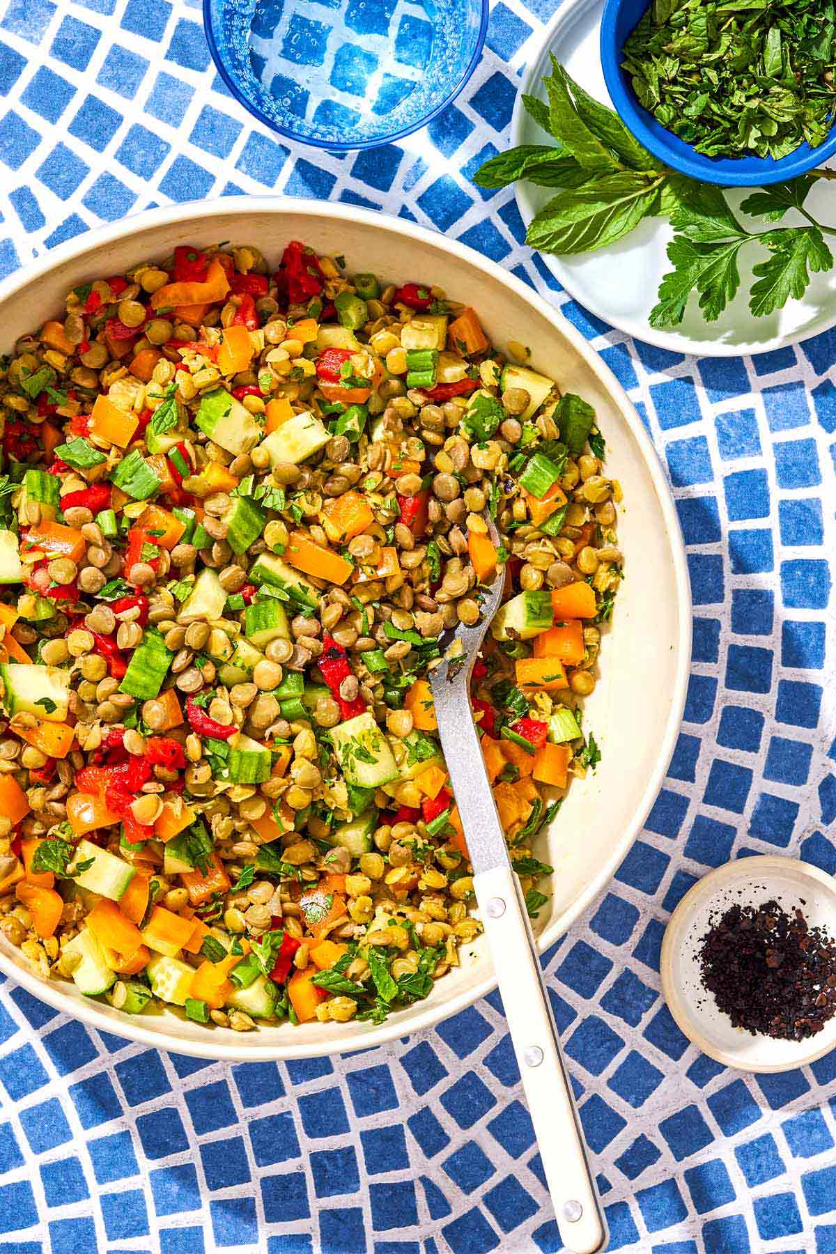 Lentil salad in a serving bowl with a spoon next to this is a glass of water, a small bowl of urfa biber, and a small plate with mint leaves and a bowl of chopped parsley.