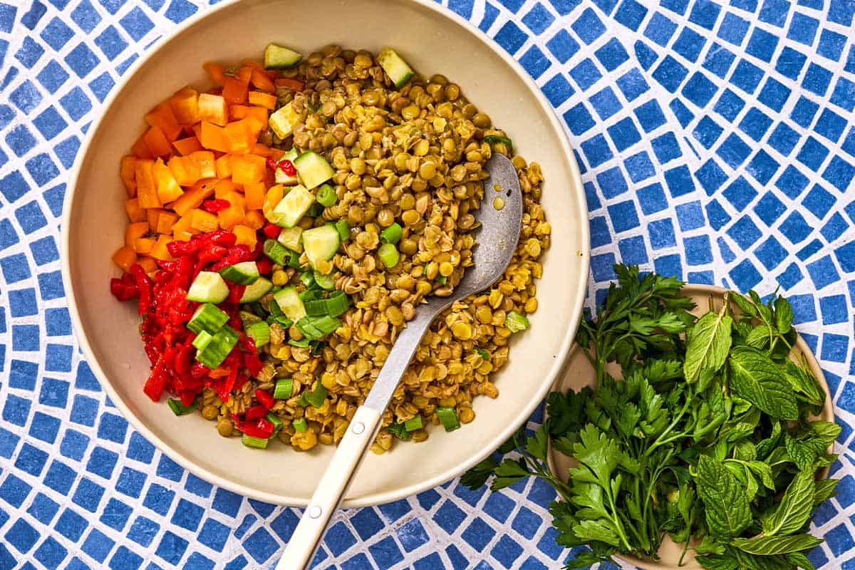 The ingredients for the lentil salad in a bowl with a spoon, just before being mixed together. Next to this is a plate with parsley and mint.