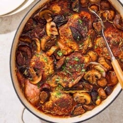 An overhead photo of a pot of wine braised chicken thighs with a serving spoon. Next to this is a stack of 2 plates.