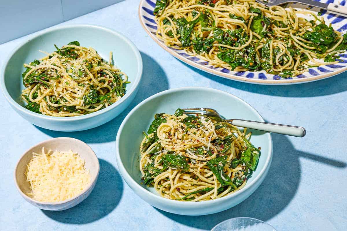 Two bowls of the za'atar garlic spinach pasta, one with a fork. Next to this is a platter of the pasta and a small bowl of parmesan cheese.