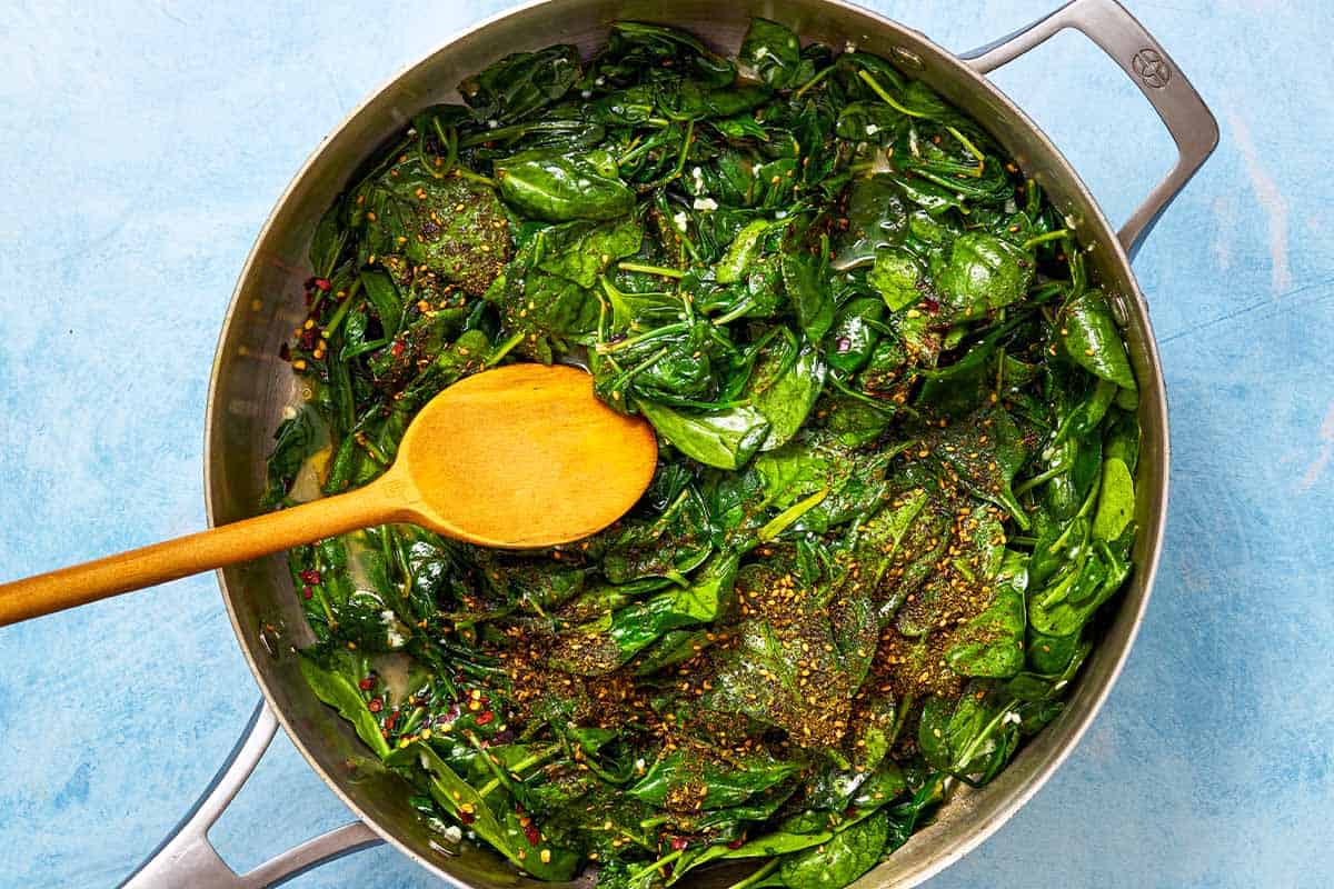 Spinach and spices being sauteed in pan with a wooden spoon.