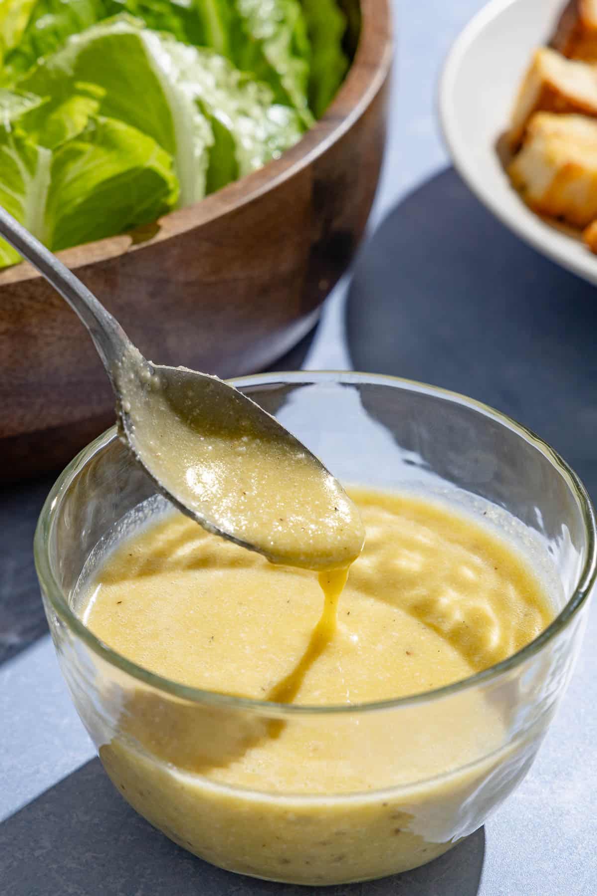 A close up of caesar salad dressing in a bowl with a spoon in front of a bowl of romaine lettuce and a plate of homemade croutons.