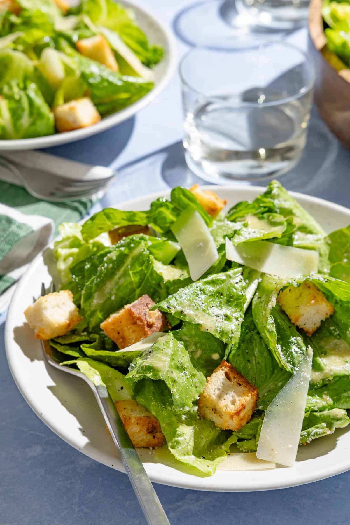 A close up of a plate of caesar salad with a fork. Next to this is another plate of the salad, a glass of water, and a fork on a cloth napkin.
