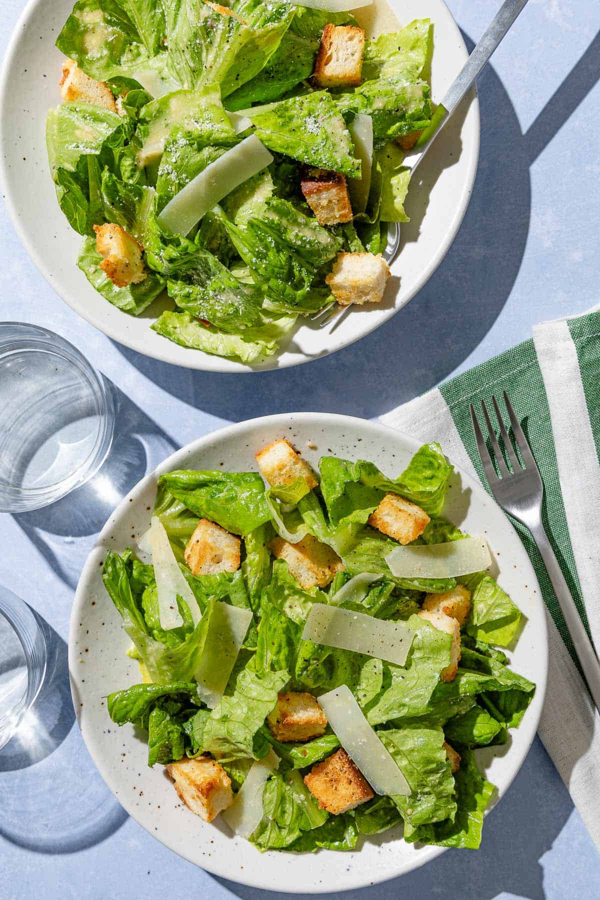 An overhead photo of 2 servings of caesar salad on plates with forks. Next to this is a cloth napkin and 2 glasses of water.