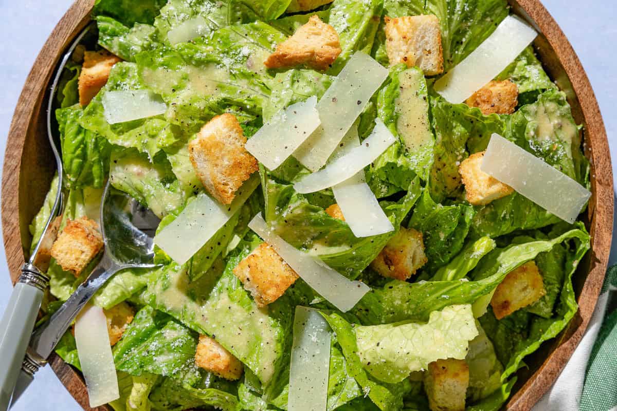 An overhead, close up photo of caesar salad in a serving bowl with serving utensils.