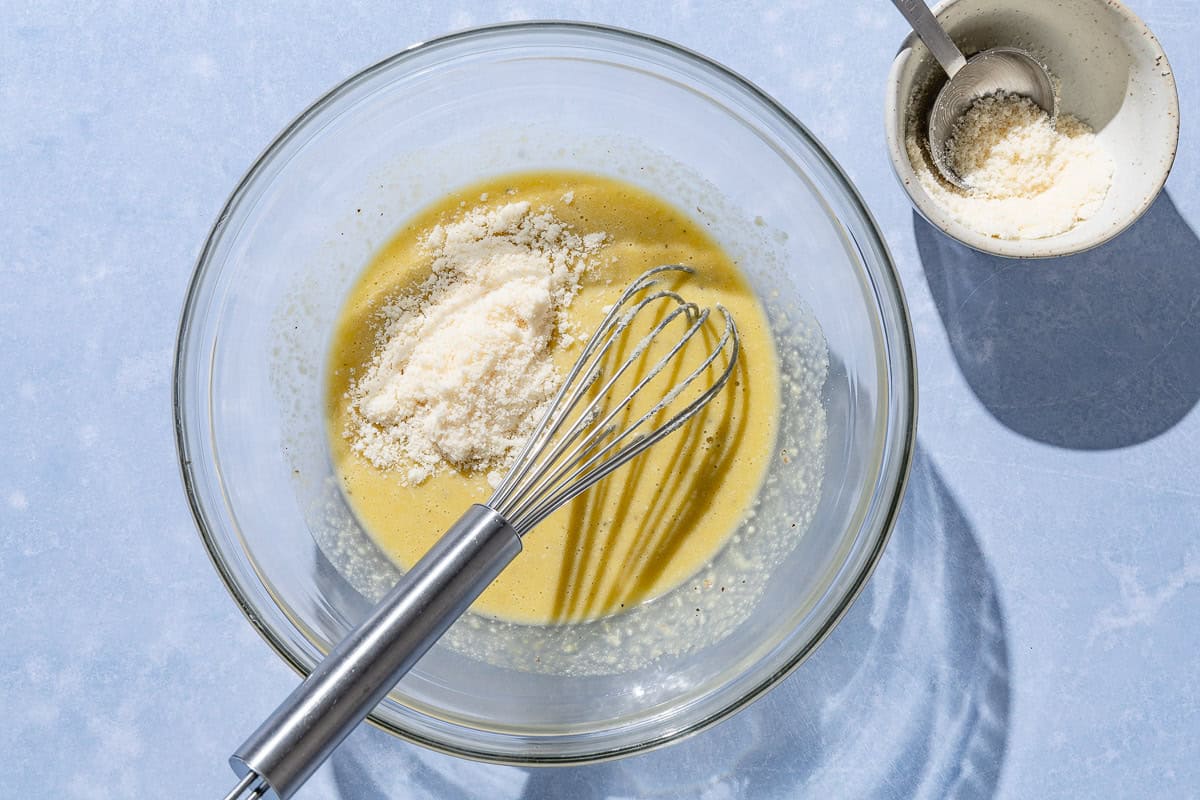 The ingredients for the caesar salad in a bowl with a whisk right after some parmesan cheese was added. Next to this is a small bowl of grated parmesan cheese with a spoon.