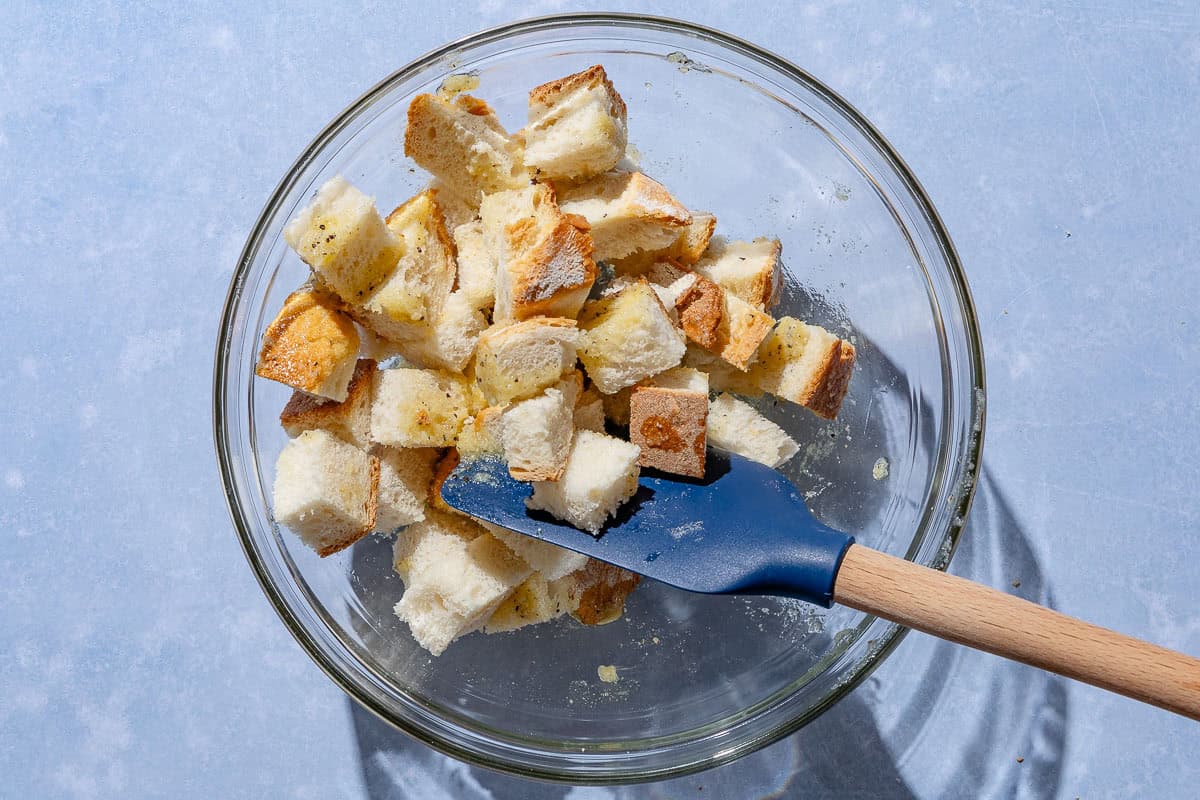 Cubes of unbaked bread being mixed with olive oil and seasonings in a bowl with a rubber spatula.