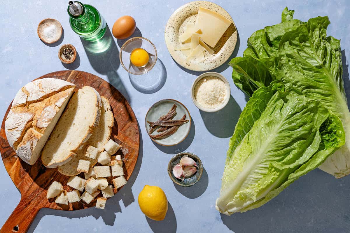 Ingredients for caesar salad including romaine lettuce, parmesan cheese, olive oil, parmesan cheese, garlic, salt, black pepper, crusty bread, anchovies, an egg, and lemon.