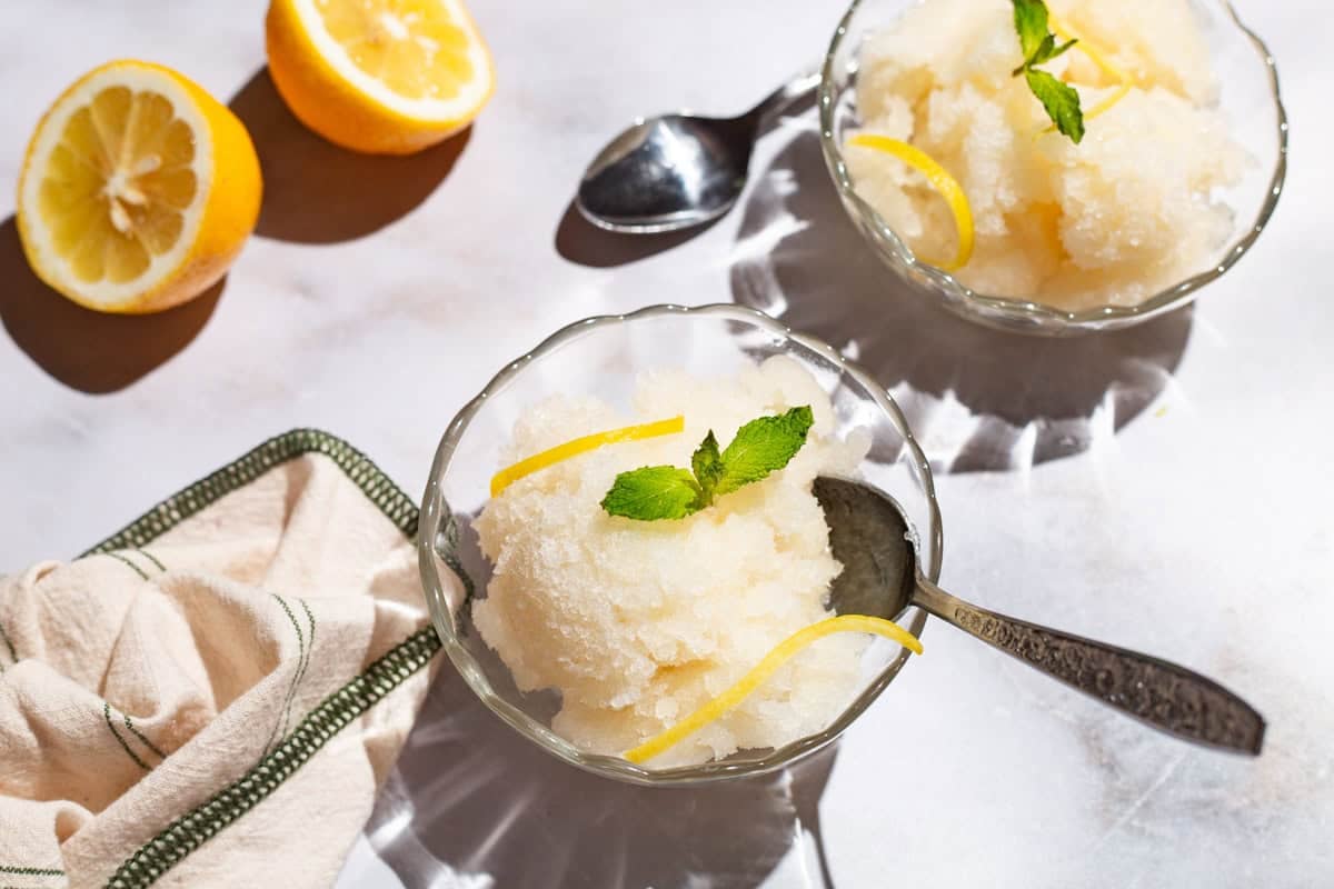 Two bowls of lemon sorbet, one with a spoon, and one next to a spoon. Next to these are 2 lemon wheels and a kitchen towel.
