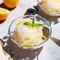 Two bowls of lemon sorbet, one with a spoon, and one next to a spoon. Next to these are 2 lemon wheels and a kitchen towel.