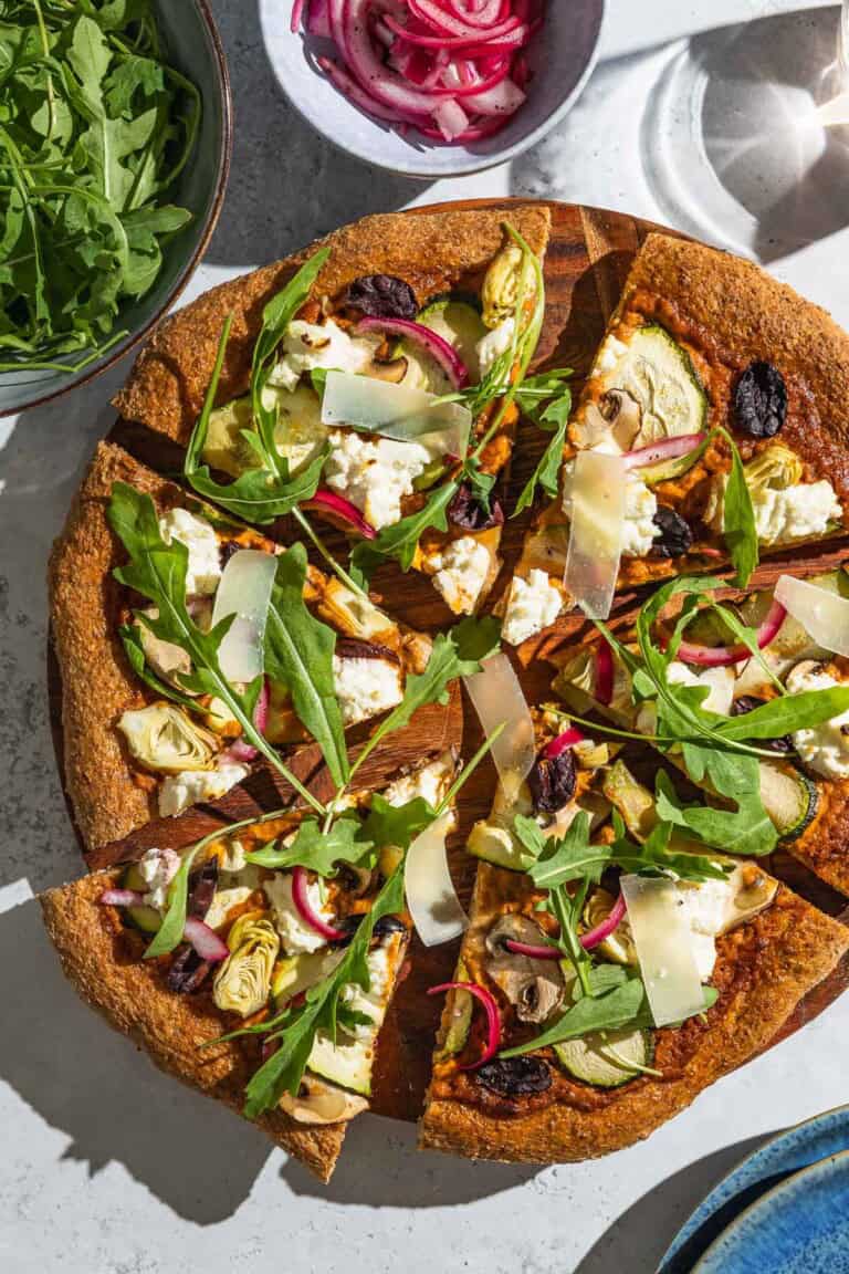 An overhead photo of a sliced vegetarian pizza on a wooden serving platter next to bowls of arugula and pickled onions.