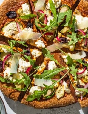 An overhead photo of a sliced vegetarian pizza on a wooden serving platter next to bowls of arugula and pickled onions.