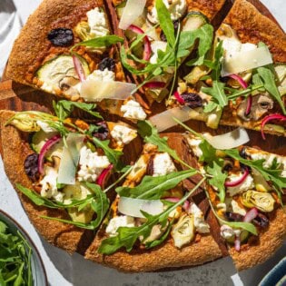 An overhead photo of a sliced vegetarian pizza on a wooden serving platter next to bowls of arugula and pickled onions.