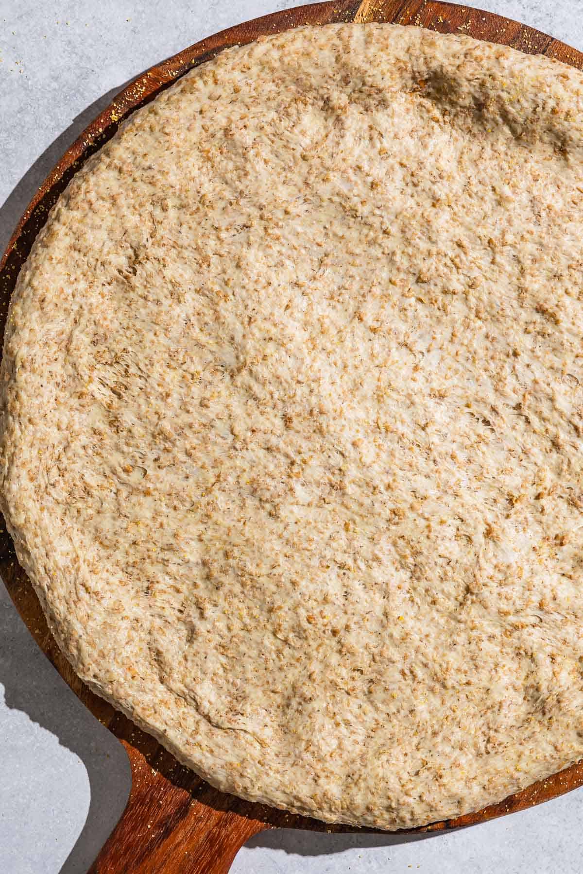 An unbaked whole wheat pizza crust on a wooden serving platter.