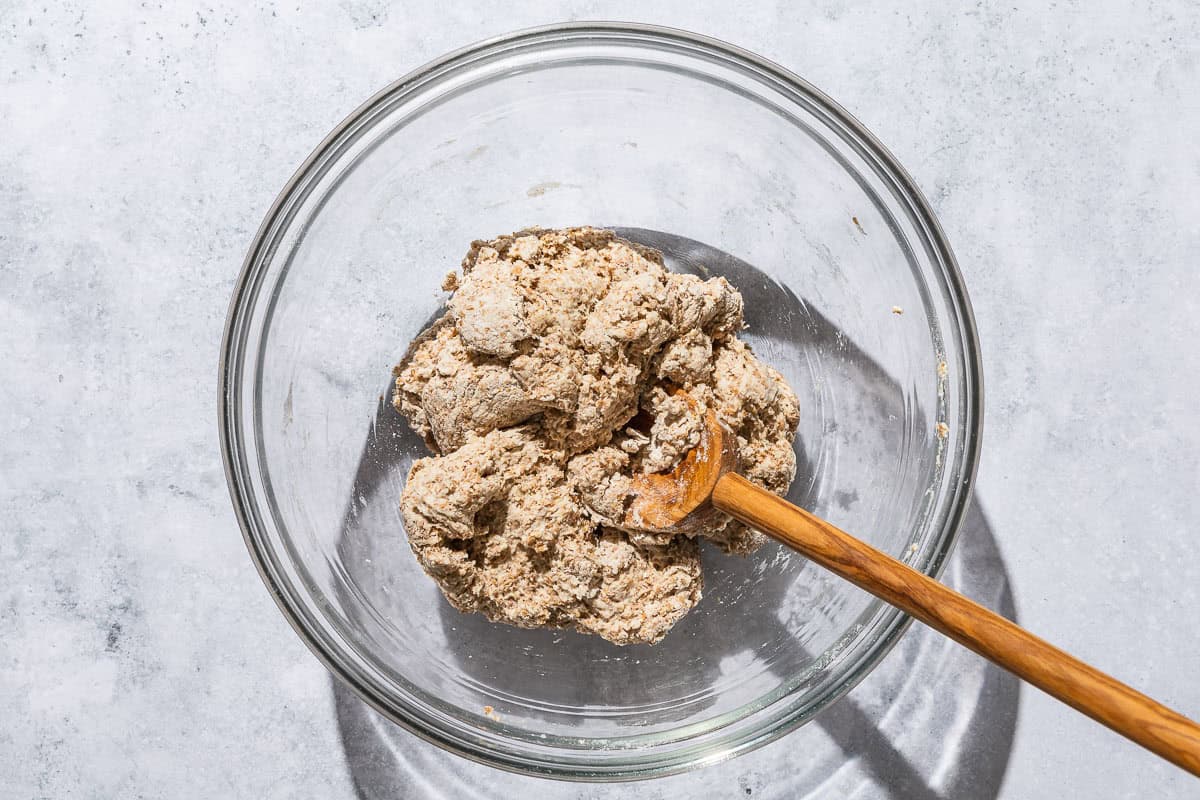 Whole wheat pizza dough being stirred in a bowl with a wooden spoon.