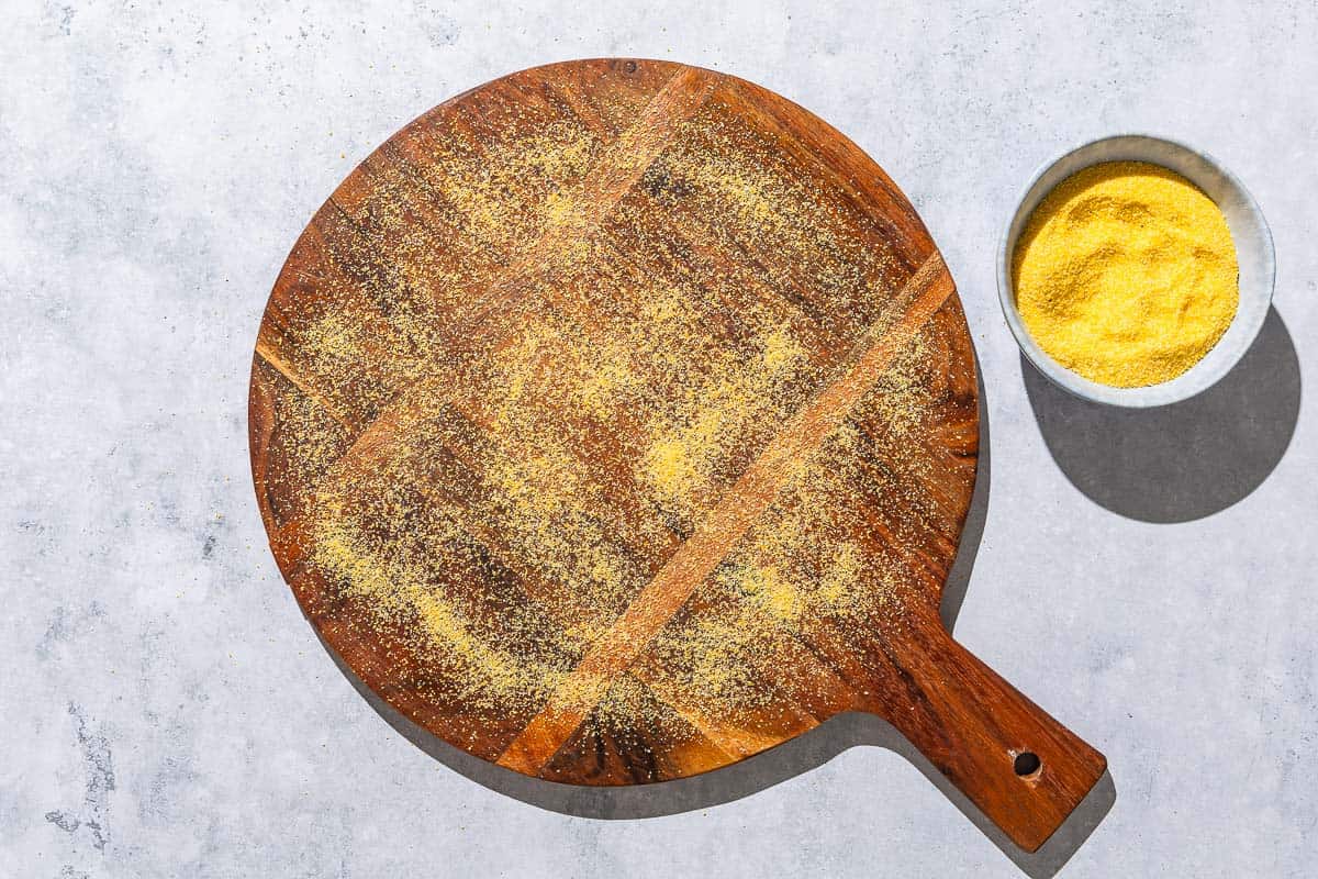An overhead photo of a wooden serving platter dusted with cornmeal next to a bowl of cornmeal.