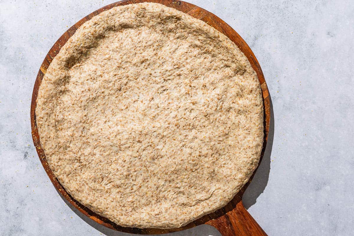 An unbaked whole wheat pizza crust on a wooden serving platter.